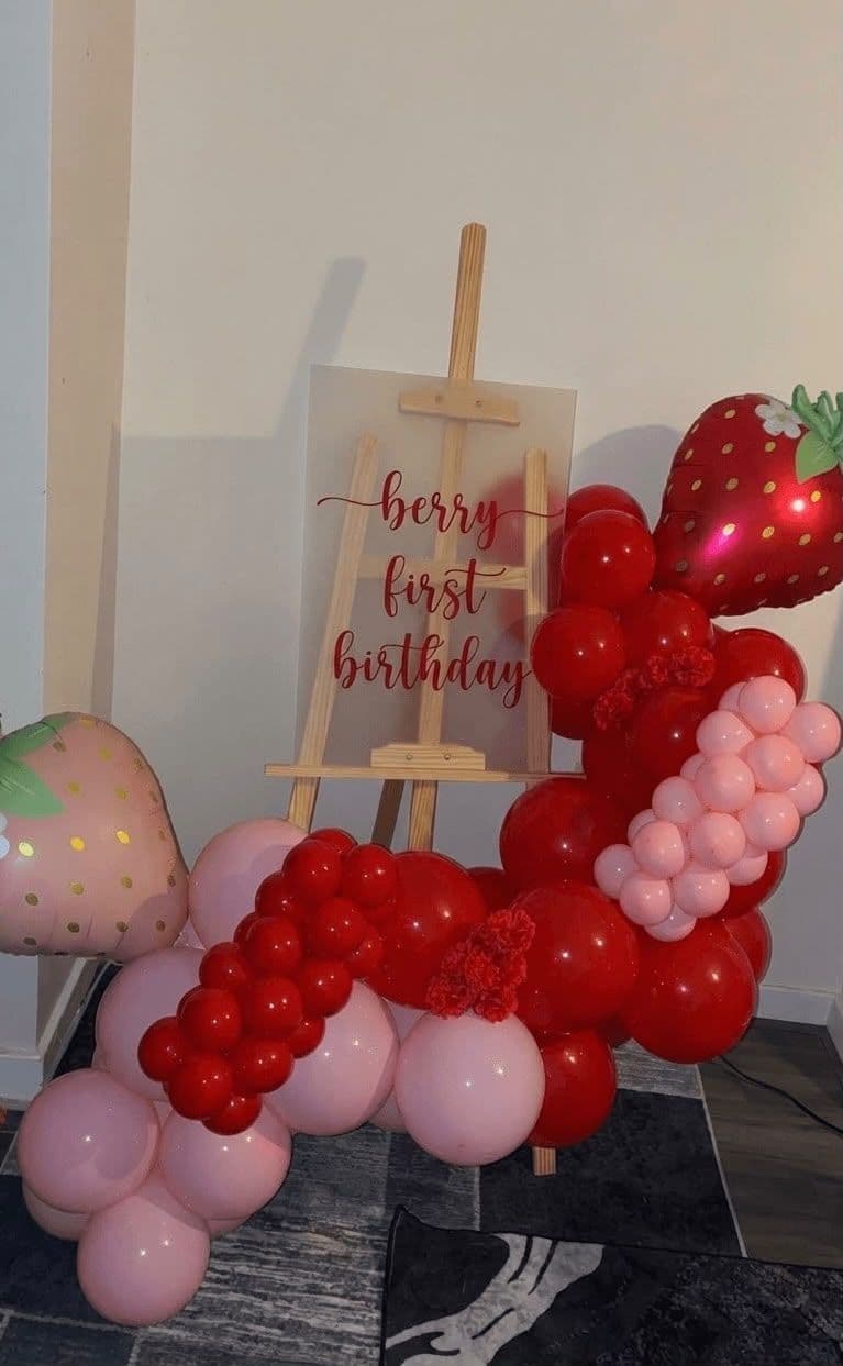 A strawberry-themed first birthday display in red and pink balloons around a "berry first birthday" sign on a wooden easel