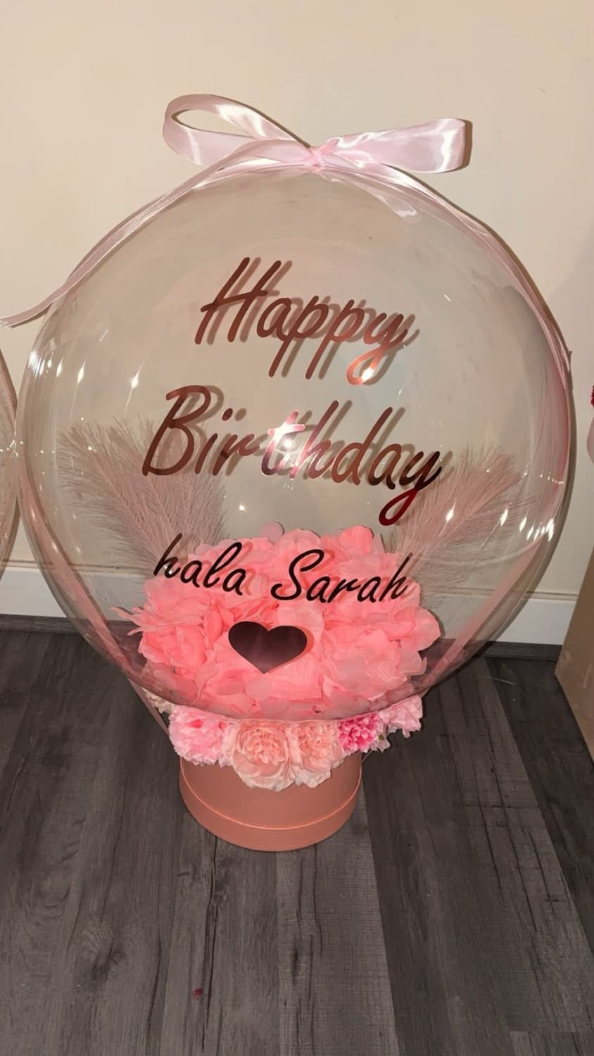 A clear bubble balloon reading Happy Birthday hala Sarah in rose gold, over a box of pink flowers