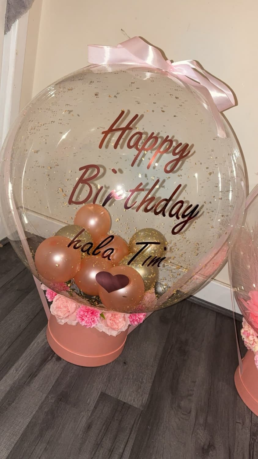 A clear glitter bubble balloon reading Happy Birthday hala Tim, filled with rose gold balloons