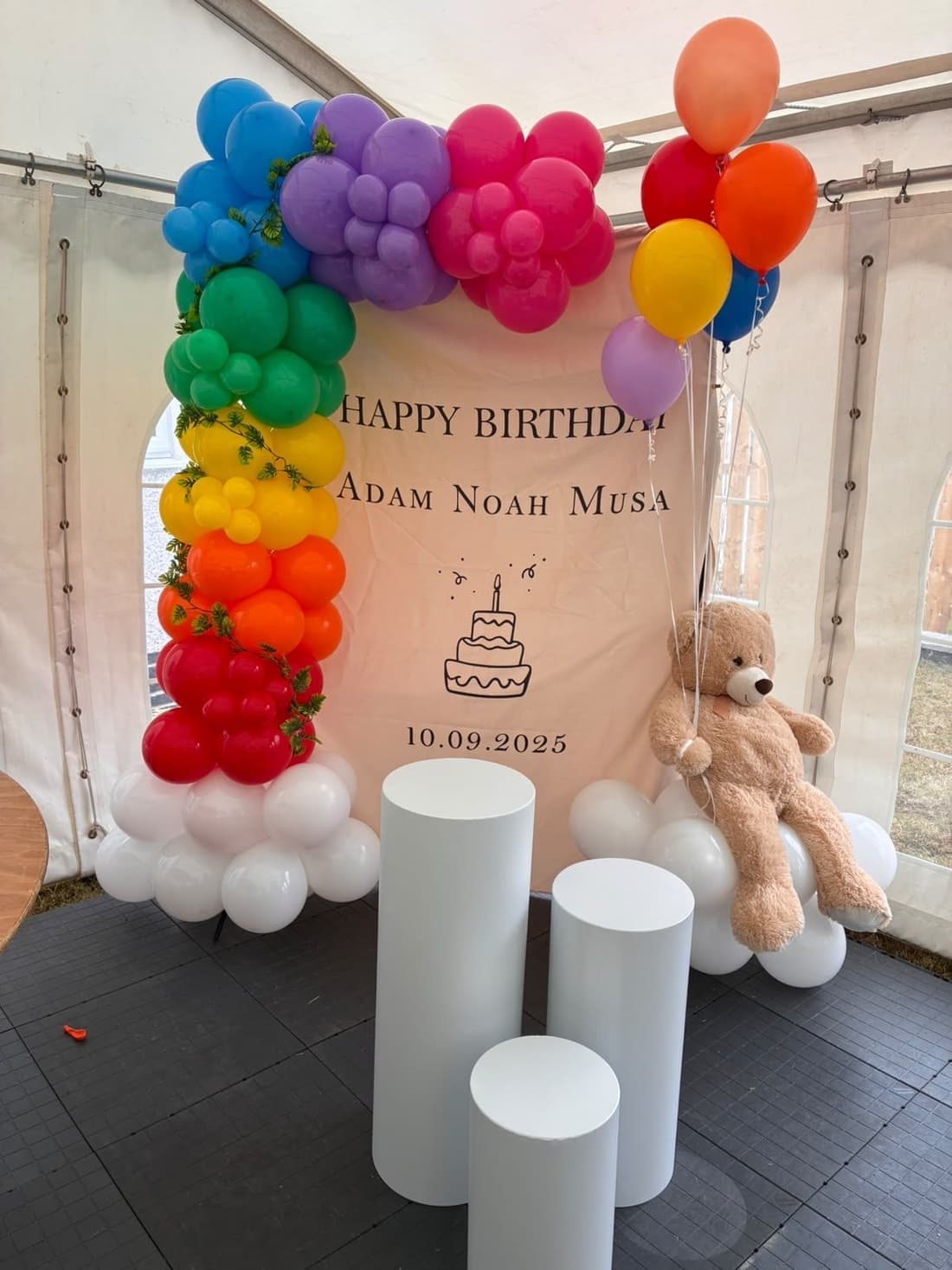 A first birthday setup with a rainbow balloon arch, a printed backdrop, white plinths and a teddy bear