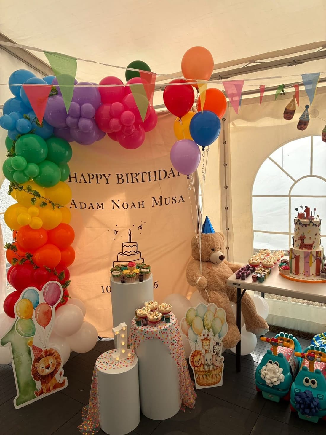 A first birthday party in a marquee with a rainbow balloon garland, bunting, a cake table and a teddy bear