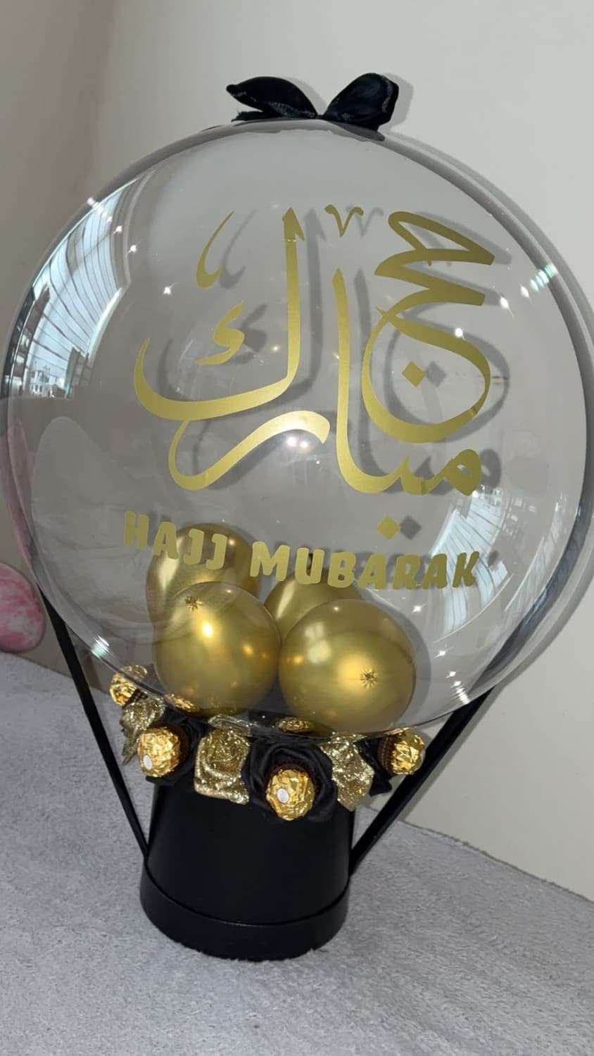 A clear bubble balloon with Hajj Mubarak in gold Arabic and English, filled with gold balloons